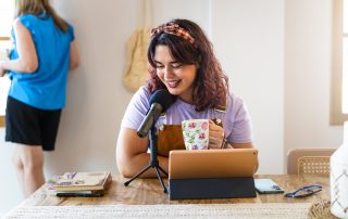 An einem Tisch sitzt eine Frau mit Mikrofon und Tablet. Sie hält eine Tasse Kaffee und lächelt. Im hinteren linken Bildrand steht eine Frau mit dem Rücken zur Kamera.